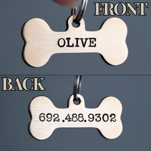 K&ouml;nnte beinhalten: Ein bronzefarbener Hundeanh&auml;nger in Knochenform. Auf der Vorderseite des Anh&auml;ngers steht "OLIVE". Auf der R&uuml;ckseite steht die Telefonnummer "692.488.9302". Der Anh&auml;nger ist an einem Metallring befestigt.
