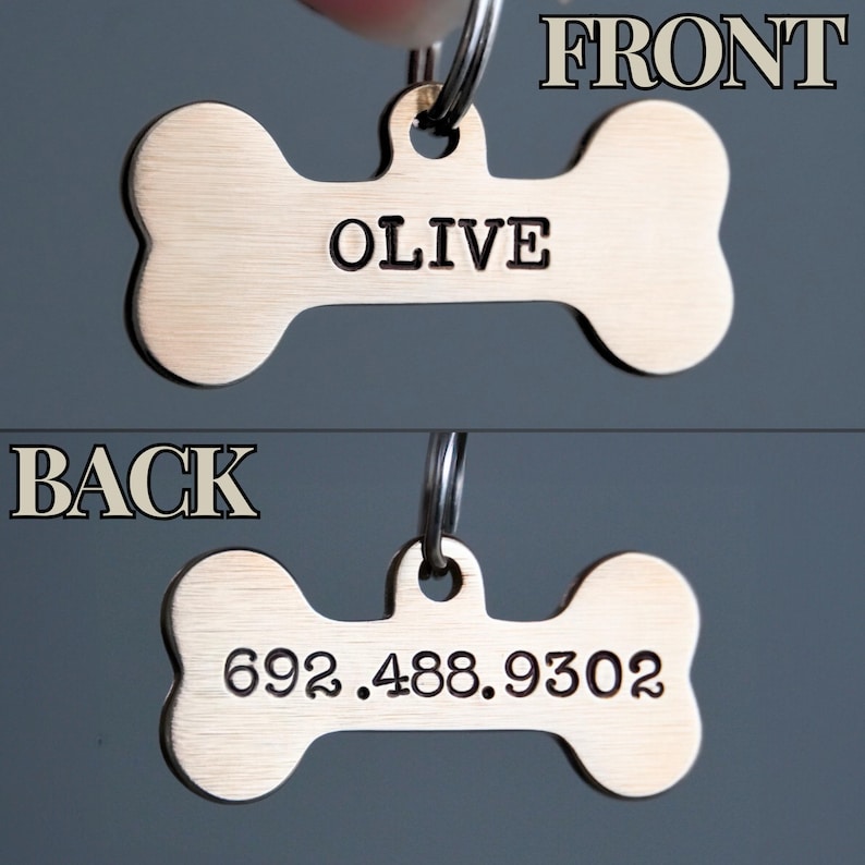 K&ouml;nnte beinhalten: Ein bronzefarbener Hundeanh&auml;nger in Knochenform. Auf der Vorderseite des Anh&auml;ngers steht "OLIVE". Auf der R&uuml;ckseite steht die Telefonnummer "692.488.9302". Der Anh&auml;nger ist an einem Metallring befestigt.