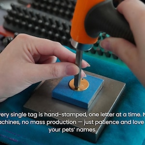 May include: A circular gold-colored metal pet tag being hand-stamped with a metal punch tool and an orange-handled hammer. The tag is secured on a square metal block with blue tape. Text reads: Every single tag is hand-stamped, one letter at a time.