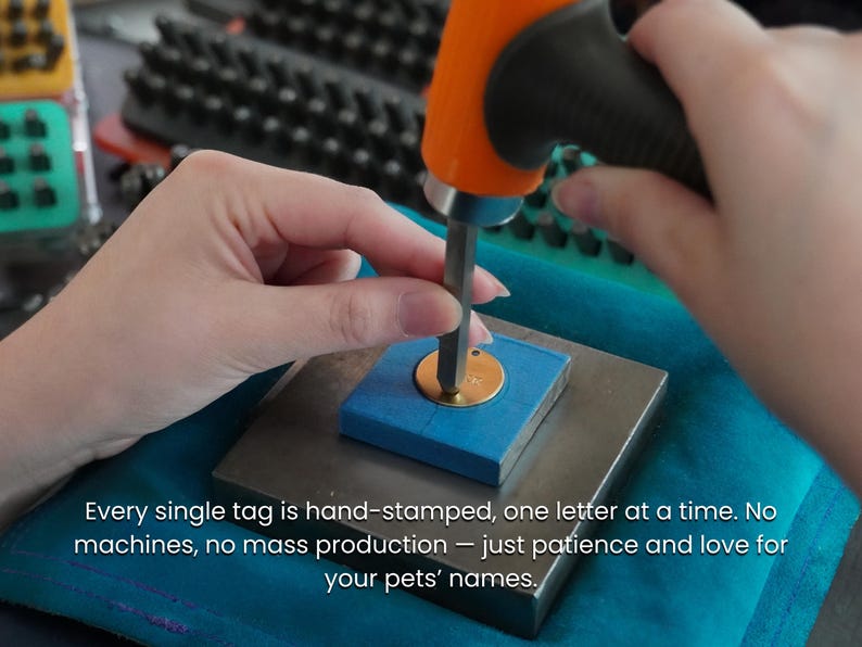 May include: A circular gold-colored metal pet tag being hand-stamped with a metal punch tool and an orange-handled hammer. The tag is secured on a square metal block with blue tape. Text reads: Every single tag is hand-stamped, one letter at a time.
