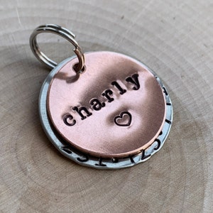 May include: A circular pet identification tag featuring a smaller copper-colored disc layered over a larger silver-colored disc. The copper disc is stamped with the name Charly and a small heart symbol. A metal split ring is attached to the top.