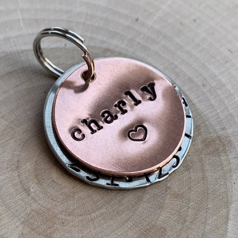 May include: A circular pet identification tag featuring a smaller copper-colored disc layered over a larger silver-colored disc. The copper disc is stamped with the name Charly and a small heart symbol. A metal split ring is attached to the top.