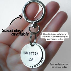 May include: A silver-toned metal keychain featuring a circular swivel clasp and two round metal tags. The top tag is stamped with the name WINSTON and a small planet icon. The bottom tag displays the numbers 236 0815 in an Uppercase Indigo font.
