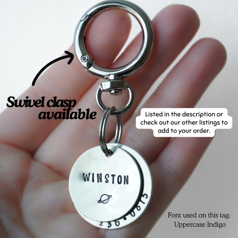 May include: A silver-toned metal keychain featuring a circular swivel clasp and two round metal tags. The top tag is stamped with the name WINSTON and a small planet icon. The bottom tag displays the numbers 236 0815 in an Uppercase Indigo font.
