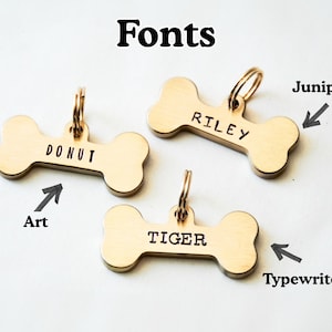 K&ouml;nnte beinhalten: Drei goldfarbene Hundeanh&auml;nger in Knochenform mit verschiedenen Schriftarten. Die Anh&auml;nger sind mit den Namen "DONUT", "RILEY" und "TIGER" graviert. Das Bild enth&auml;lt auch das Wort "Fonts" oben.