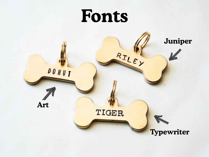 K&ouml;nnte beinhalten: Drei goldfarbene Hundeanh&auml;nger in Knochenform mit verschiedenen Schriftarten. Die Anh&auml;nger sind mit den Namen "DONUT", "RILEY" und "TIGER" graviert. Das Bild enth&auml;lt auch das Wort "Fonts" oben.