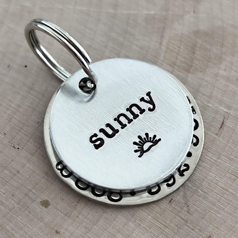 May include: A circular silver-toned metal pet identification tag featuring a brushed finish. The top layer is engraved with the name sunny and a small sun icon. A larger base disc is visible underneath with a decorative stamped border pattern.