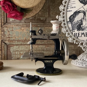 May include: A vintage black Singer sewing machine with a silver needle and foot pedal. The machine is on a white surface with a wooden base and a metal clamp.