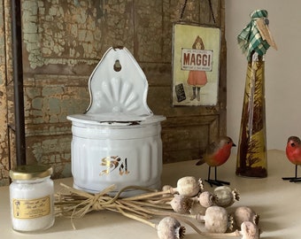 May include: White enamel salt box with a gold inscription "Sel" hanging on a wall. The salt box is next to a glass jar of Fleur de Sel, a small bottle with a green and white checkered top, and a bouquet of dried poppy seed pods.