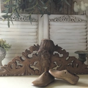 Può includere: Un pezzo decorativo in legno intagliato con dettagli intricati, che ricorda un motivo floreale. Due forme di scarpe in legno sono posizionate davanti al pezzo, una è marrone e l'altra è di una tonalità di marrone più chiara.