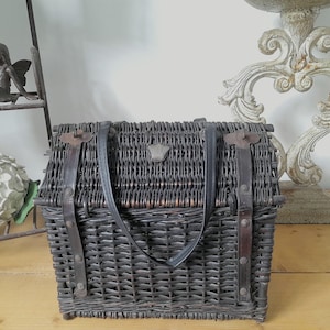 May include: A dark gray wicker picnic basket with a leather handle and straps. The basket has a rectangular shape with a hinged lid and metal clasps. A small decorative emblem is centered on the front.