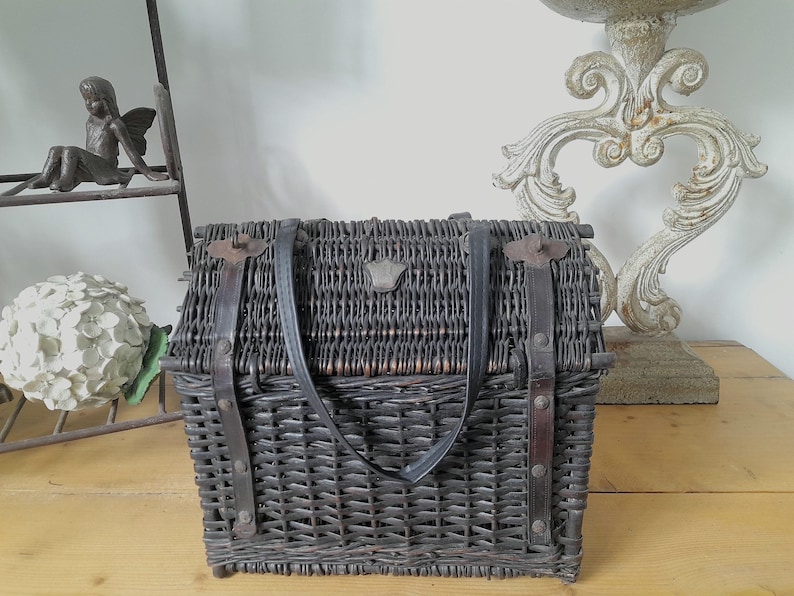 May include: A dark gray wicker picnic basket with a leather handle and straps. The basket has a rectangular shape with a hinged lid and metal clasps. A small decorative emblem is centered on the front.
