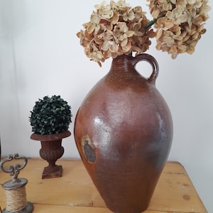Op de afbeelding: Een grote, bruine keramische kruik met een handvat, gevuld met gedroogde beige bloemen. De kruik heeft een afgeronde body en een smalle hals. Een kleine topiary en een decoratieve spoel zijn ook zichtbaar.