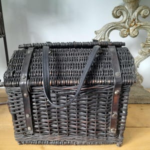 May include: A dark brown wicker picnic basket with a rectangular shape and a hinged lid. The basket features black leather straps and a handle for carrying. The basket is designed with a woven pattern.