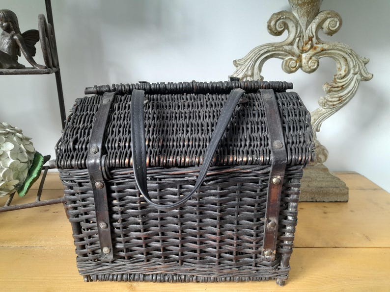 May include: A dark brown wicker picnic basket with a rectangular shape and a hinged lid. The basket features black leather straps and a handle for carrying. The basket is designed with a woven pattern.