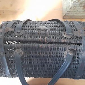 May include: A dark gray, woven wicker basket with black leather straps and handles. The basket has a rectangular shape with a hinged lid and metal clasp. The handles are curved and attached to the sides.