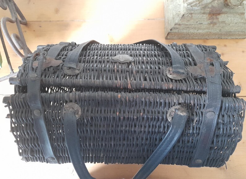May include: A dark gray, woven wicker basket with black leather straps and handles. The basket has a rectangular shape with a hinged lid and metal clasp. The handles are curved and attached to the sides.