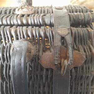 May include: Close-up of a vintage wicker basket with a dark finish. The basket features woven construction and leather straps with metal clasps. The handle is visible at the top. The basket is likely used for picnics or storage.