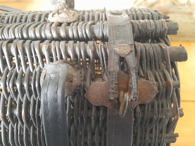 May include: Close-up of a vintage wicker basket with a dark finish. The basket features woven construction and leather straps with metal clasps. The handle is visible at the top. The basket is likely used for picnics or storage.