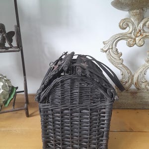 May include: A dark gray woven wicker basket with a house-like shape. The basket has a rectangular base and a curved roof, with a handle on top. The wicker is tightly woven, creating a textured surface. The basket is likely used for storage or decoration.