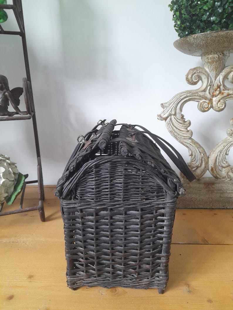 May include: A dark gray woven wicker basket with a house-like shape. The basket has a rectangular base and a curved roof, with a handle on top. The wicker is tightly woven, creating a textured surface. The basket is likely used for storage or decoration.