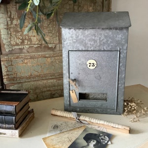 Pu&ograve; includere: Una cassetta postale in metallo in stile vintage con una finitura grigia invecchiata. La cassetta postale ha un'apertura rettangolare con uno sportello e un piccolo numero in ottone "73" sul davanti.