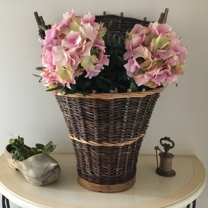 May include: A large, dark brown woven basket filled with pink and green hydrangeas. A wooden clog planter with greenery sits to the left. A small spool and metal object are on the table.