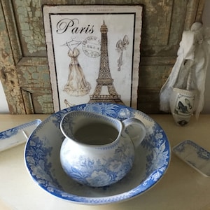 Könnte beinhalten: Ein blaues und weißes Porzellan-Waschbecken und -Krug-Set mit einem Blumenmuster. Das Set befindet sich auf einer weißen Oberfläche mit einem Schild mit der Aufschrift "Paris" im Hintergrund.