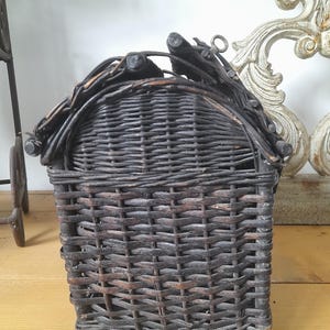 May include: A dark brown woven wicker basket with a curved top and a handle. The basket is rectangular with a square base and is made of tightly woven wicker. The handle is made of twigs and branches. The basket is on a light brown wooden surface.
