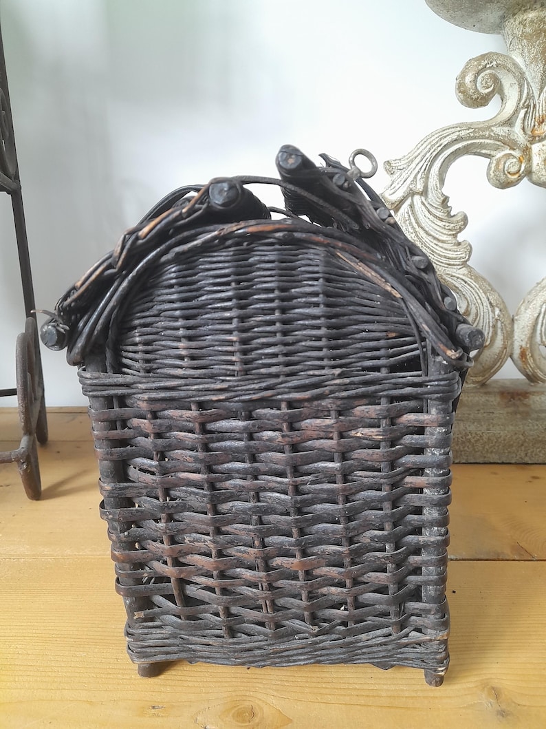 May include: A dark brown woven wicker basket with a curved top and a handle. The basket is rectangular with a square base and is made of tightly woven wicker. The handle is made of twigs and branches. The basket is on a light brown wooden surface.