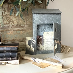 Pu&ograve; includere: Una cassetta postale vintage in metallo galvanizzato con una porta a cerniera e una piccola finestra. La cassetta postale &egrave; aperta e contiene una lettera scritta a mano e una fotografia vintage di una donna con un cappello. La cassetta postale &egrave; appoggiata su un tavolo con un rotolo di carta e una pila di libri.