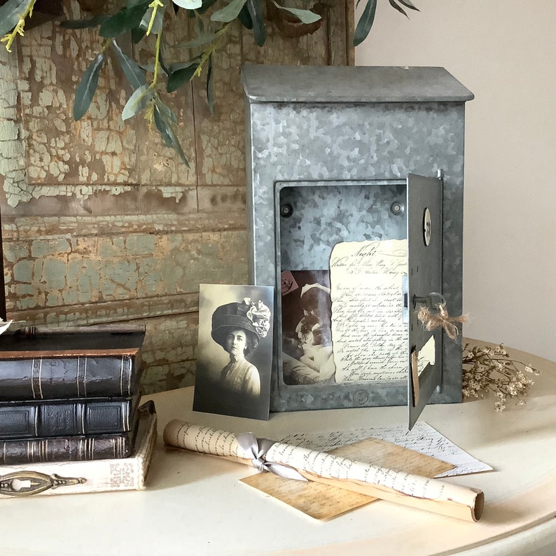 Pu&ograve; includere: Una cassetta postale vintage in metallo galvanizzato con una porta a cerniera e una piccola finestra. La cassetta postale &egrave; aperta e contiene una lettera scritta a mano e una fotografia vintage di una donna con un cappello. La cassetta postale &egrave; appoggiata su un tavolo con un rotolo di carta e una pila di libri.