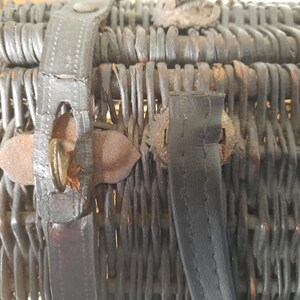 May include: Close-up of a vintage wicker basket with dark brown woven construction. The basket features leather straps and metal clasps for closure. The basket's aged appearance suggests it is an antique.