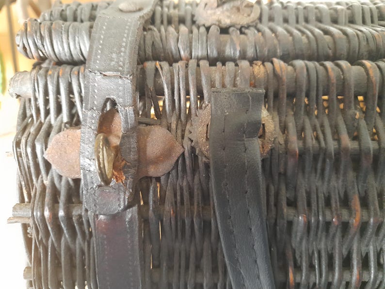 May include: Close-up of a vintage wicker basket with dark brown woven construction. The basket features leather straps and metal clasps for closure. The basket's aged appearance suggests it is an antique.