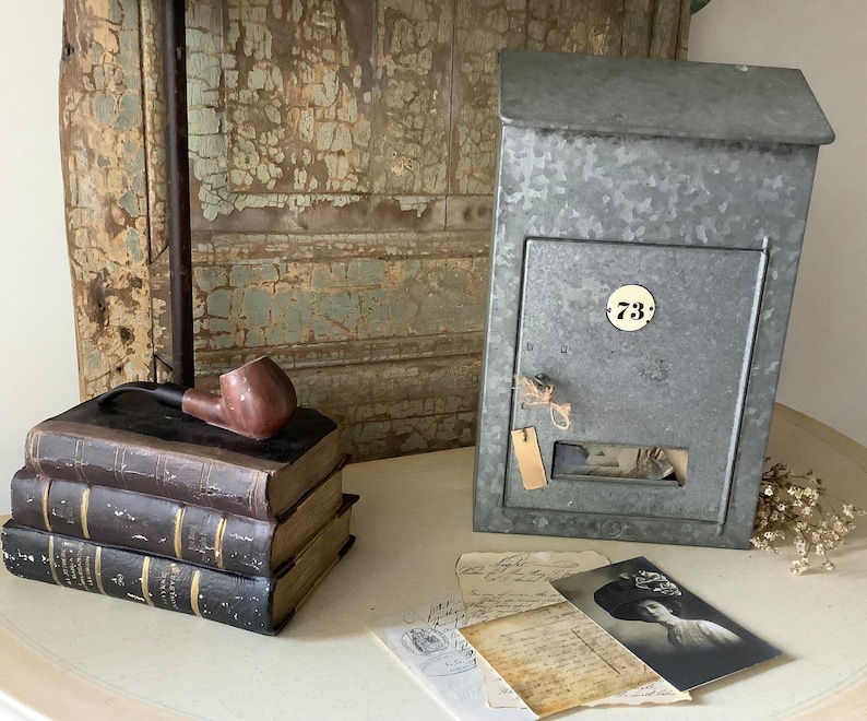 Pu&ograve; includere: Una cassetta postale vintage in metallo zincato con il numero 73 sul davanti. La cassetta postale &egrave; appoggiata su un tavolo con una pila di libri e una pipa.