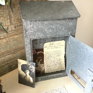Pu&ograve; includere: Una cassetta delle lettere vintage in metallo zincato con uno sportello incernierato aperto, che rivela una lettera manoscritta e una fotografia d'epoca all'interno. La cassetta delle lettere ha un aspetto rustico e invecchiato.