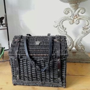 May include: A dark gray woven wicker basket with a leather handle and straps. The basket has a rectangular shape and is secured with metal clasps. A decorative metal emblem is on the front.