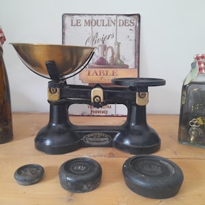 Peut inclure: Une balance ancienne noire et dorée avec un bol en laiton, accompagnée de trois poids et de deux bouteilles décoratives. Une bouteille est recouverte d'un tissu à carreaux rouges et blancs. Une enseigne en arrière-plan indique "Le Moulin des Oliviers".