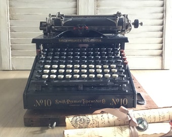 May include: A vintage black Smith Premier Typewriter No. 10 with a wooden case. The typewriter has a black metal body with a black keyboard and a black carriage return lever. The typewriter is sitting on a wooden surface with two rolled up pieces of paper.
