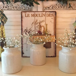 May include: Three speckled white ceramic vases with baby's breath flowers. Behind them is a vintage sign that reads "Le Moulin Des Oliviers" and two decorative bottles. The background features white shutters.