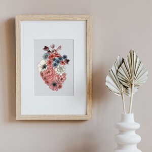 May include: A framed print of a heart-shaped arrangement of pink, white, and blue flowers. The flowers are arranged in a heart shape and are surrounded by a white background. The print is framed in a light wood frame.