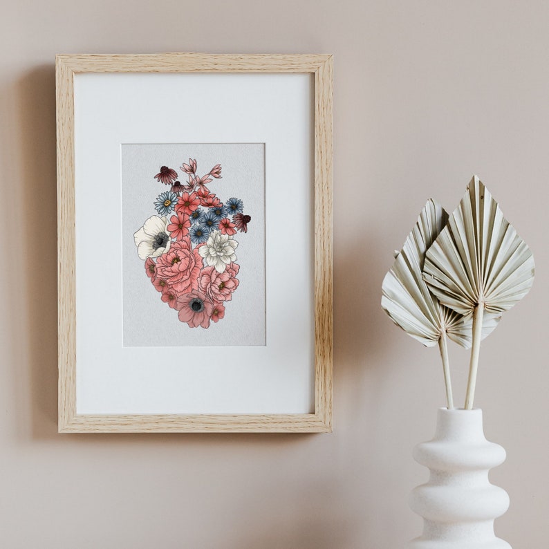 May include: A framed print of a heart-shaped arrangement of pink, white, and blue flowers. The flowers are arranged in a heart shape and are surrounded by a white background. The print is framed in a light wood frame.