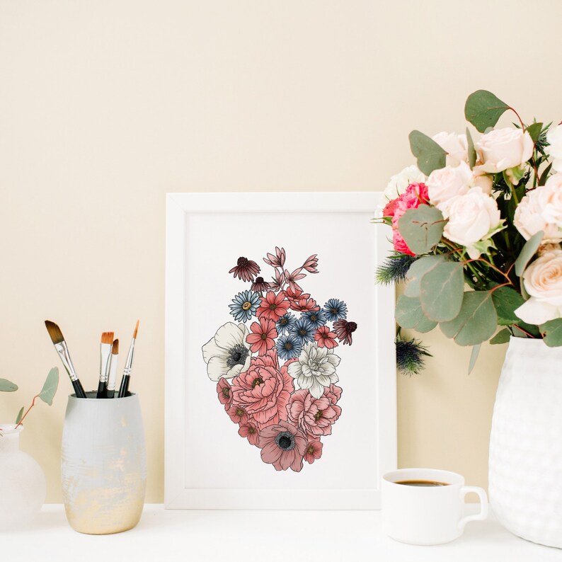 May include: A white framed print with a floral heart design. The heart is made up of pink, red, and blue flowers. The print is sitting on a table with a vase of flowers and a cup of coffee.