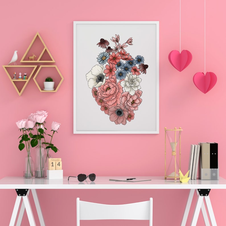 May include: A white framed print of a floral heart with pink, red, and blue flowers on a white background. The print is hanging on a pink wall with a wooden shelf and a wooden triangle shelf above it. There is a wooden hourglass, a stack of books, and a white desk with a white chair in front of it.