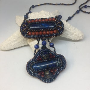 May include: A beaded necklace with a blue and red pendant featuring a lapis lazuli stone. The pendant is made of small beads and has a geometric design.