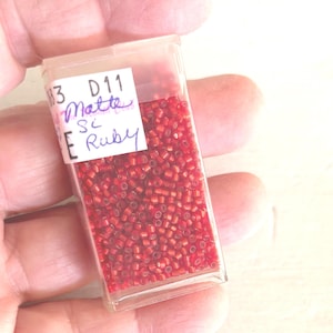 May include: A clear plastic container filled with small, matte red beads. A white label on the container reads "83 D11 Matte Si Ruby". The beads are a deep ruby color, and the container is held in a hand.