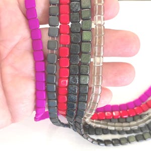 May include: Close-up of several strands of square beads in various colors, including fuchsia, gray, red, black, olive green, and clear. The beads are strung together, creating a textured and colorful display. Ideal for jewelry making.