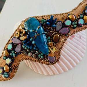 May include: A blue agate gemstone is surrounded by gold, turquoise, and purple beads on a cork base. The base is decorated with a variety of beads and stones, creating a unique and intricate design.