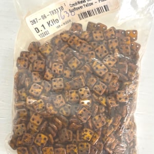 May include: A clear plastic bag filled with small, square beads. The beads are a mix of yellow and brown with black dots, resembling dice. The bag has a label with text including "CzechMates QuadraTile" and "Sunflower Yellow - Picasso".