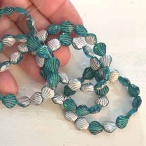 May include: Strands of iridescent teal and silver seashell-shaped beads. The beads are strung together, creating a necklace or bracelet. The beads have a textured, ridged surface, and the colors shimmer in the light. The overall theme is nautical or beach-inspired.